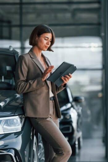 With tablet in hands. Woman is indoors near brand new automobile indoors